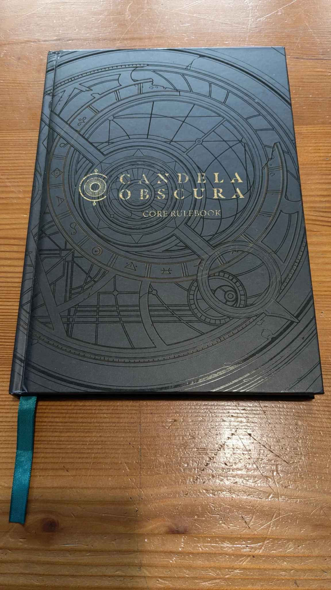 Cover of Candela Obscura. Mostly black with a light reflecting imprint of occult symbols and Candela Obscura written in the middle.