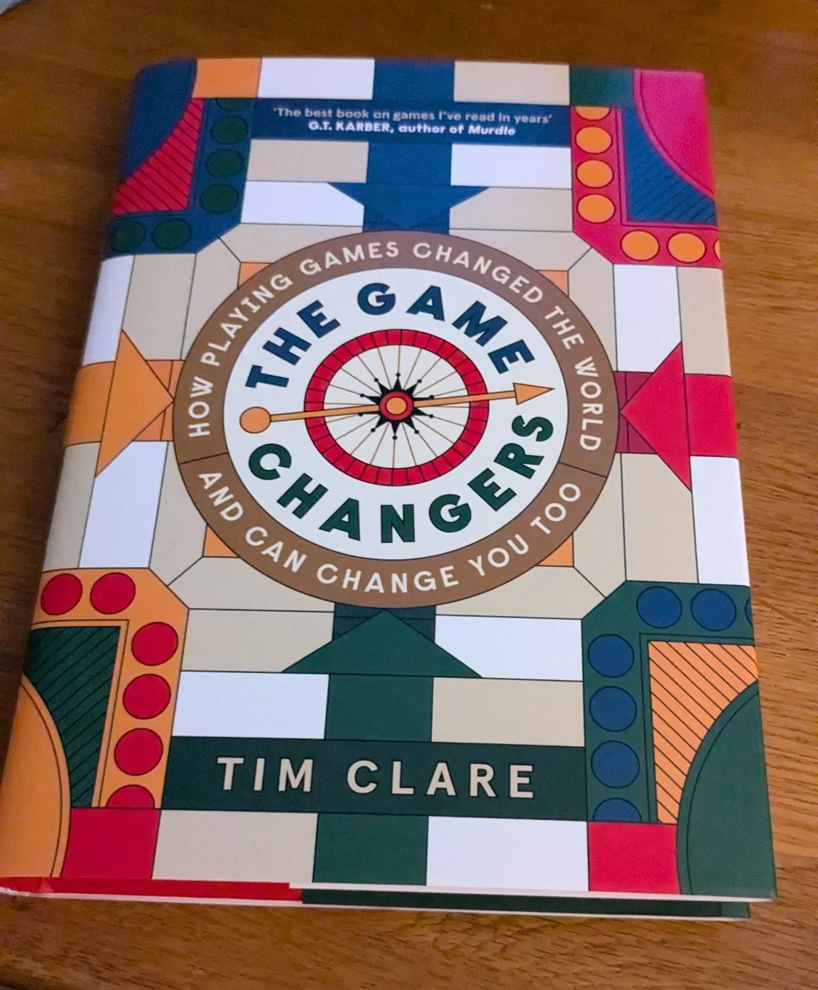 The Game Changers book lies on a table. The title in the middle of the book surrounded by a ring of words that says 'How playing games changed the world and can change you to'. The background of the book is like a ludo board.