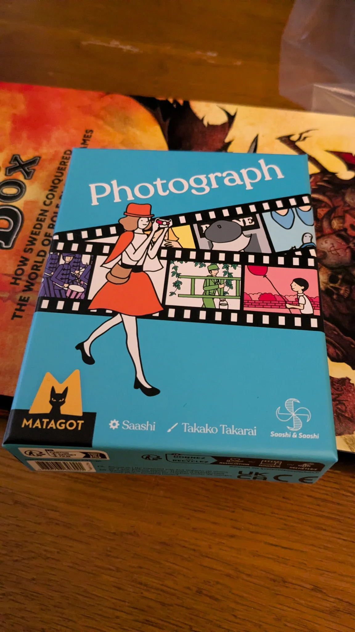 The box of photograph sits on a book. The box is blue with a film reel with various picture on it. In front of the film reel is a woman with a camera.